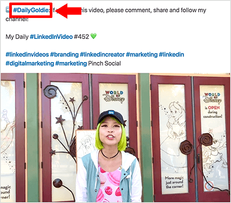 Това е екранна снимка, която илюстрира как Goldie Chan използва хаштагове в текста на своите видео публикации в LinkedIn. Червените описания сочат към хаштага #DailyGoldie в текста, който е уникален за нейните видео публикации и й помага да проследява споделянията. Публикацията включва и други подходящи хаштагове, които помагат на хората да намерят нейния видеоклип, включително #LinkedInVideo. Във видеоизображението Голди стои пред някои врати на дисплей на World of Disney. Тя е азиатка със зелена коса. Тя е с черна шапка LinkedIn, черна колие-чокър, розова риза с печат от макарон и синьо-бяло яке.